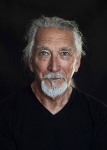 A photo from the chest up of M. John Harrison, an older white man with white hair pulled back into a ponytail and a white goatee. He is wearing black and is in front of a black background.